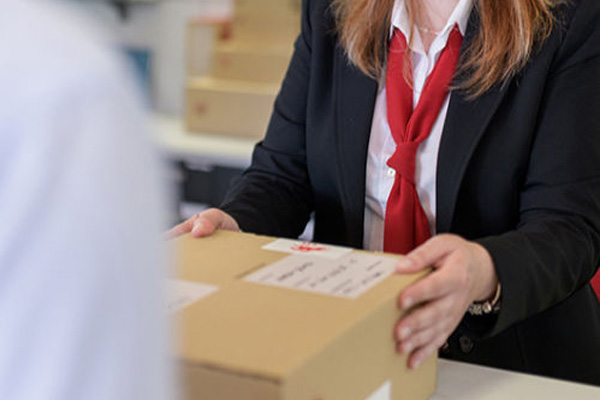 Senhora de fato com uma gravata vermelha segura numa caixa com a morada do destinatário. O remetente parece estar a entregar a caixa.
